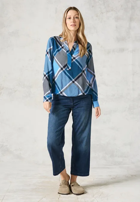 Frau in blau-karierter Bluse und weiter Jeans steht vor hellem Hintergrund.